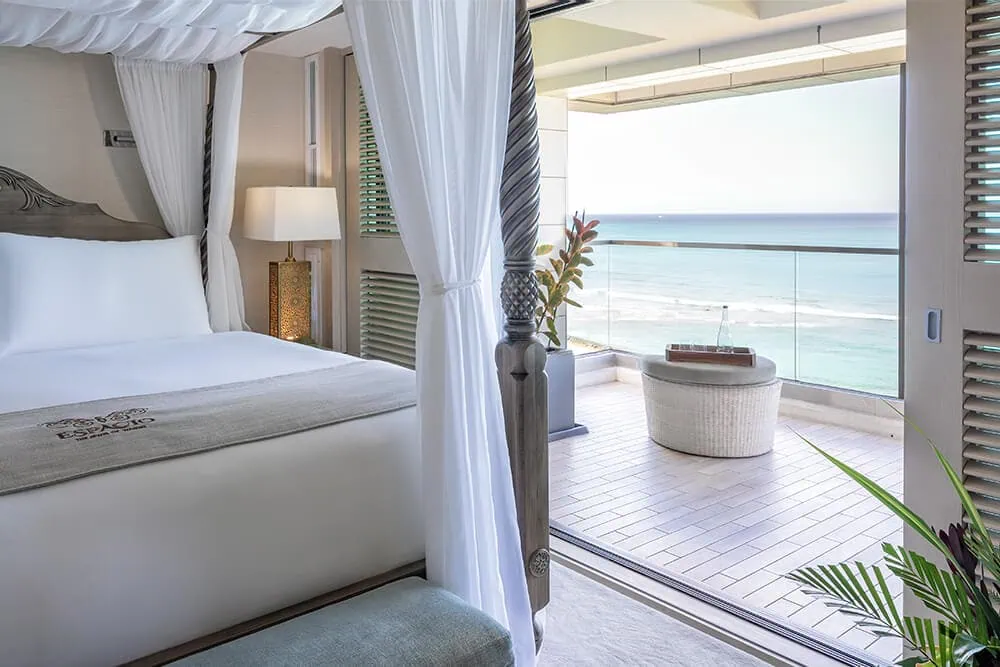 Room with a canopy bed, with a lamp and doorway to balcony with ocean view.